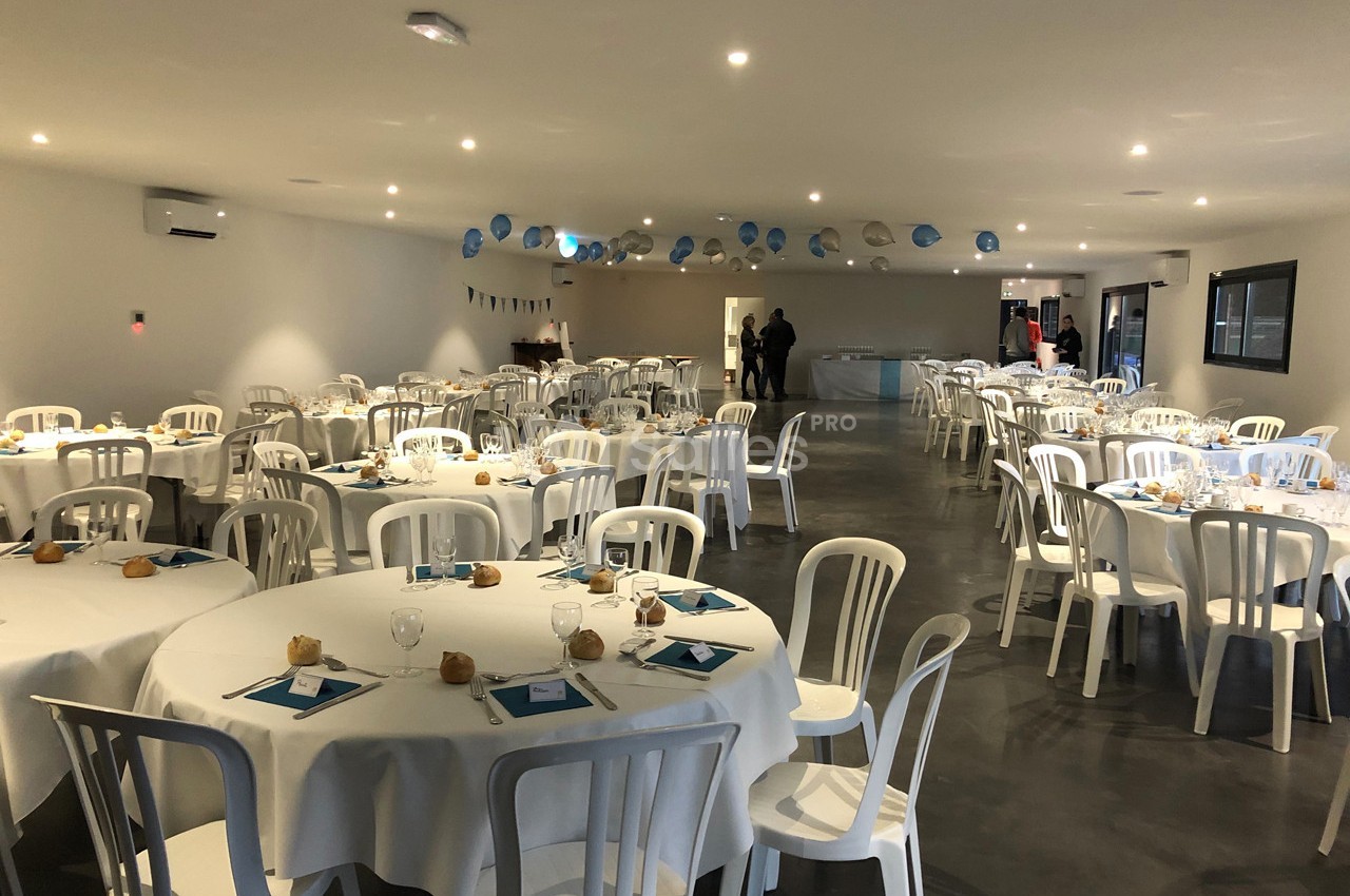 Salle de réception décorée avec des tables rondes dressées, nappes blanches et chaises en plastique blanc.