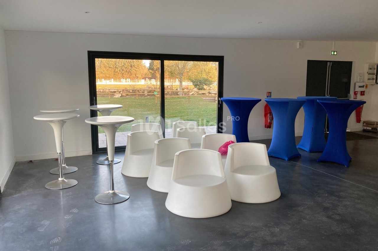 Salle lumineuse avec mobilier moderne, comprenant des tables hautes blanches et bleues et des sièges design.