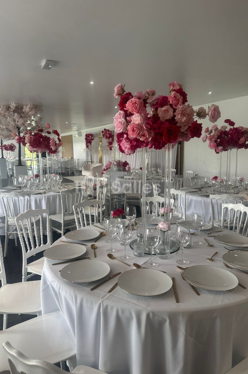 Salle de réception décorée avec des tables rondes, nappes blanches et centres de table composés de fleurs roses.