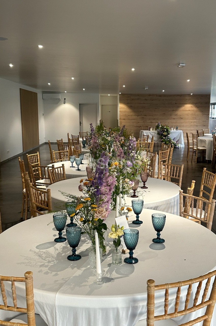 Salle de réception décorée avec des tables rondes, nappes blanches, chaises en bois et bouquets de fleurs.