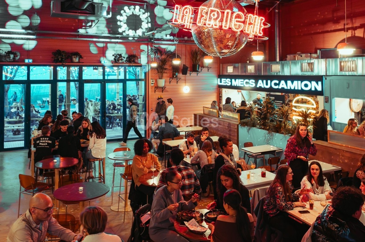 Salle animée avec des groupes de personnes dînant à des tables, sous une enseigne lumineuse ’La Friche’.
