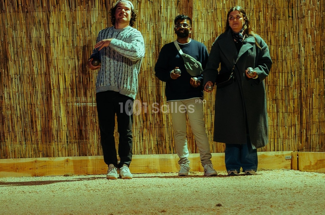 Trois personnes jouent à la pétanque sur un terrain en gravier, devant une clôture en bambou.