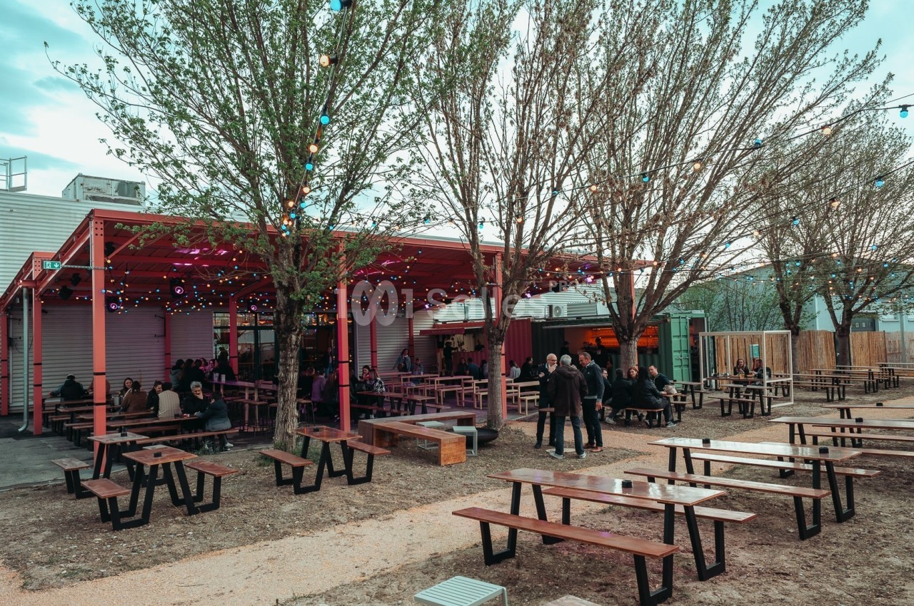Terrasse extérieure avec tables en bois, éclairages suspendus et quelques personnes assises sous un auvent rouge.