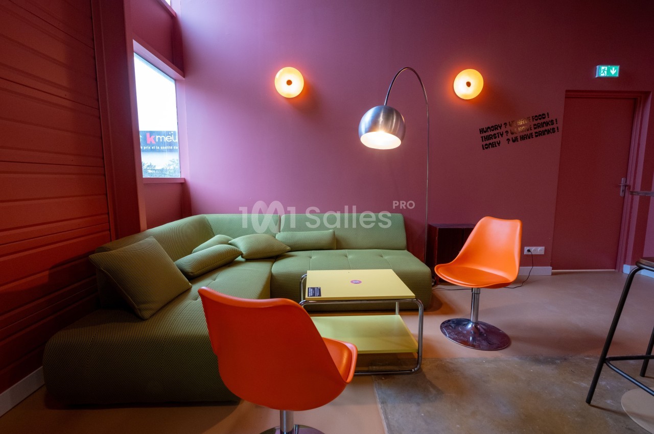 Salon moderne avec canapé vert, deux chaises orange, table basse jaune et lampadaire métallique dans une pièce aux murs…