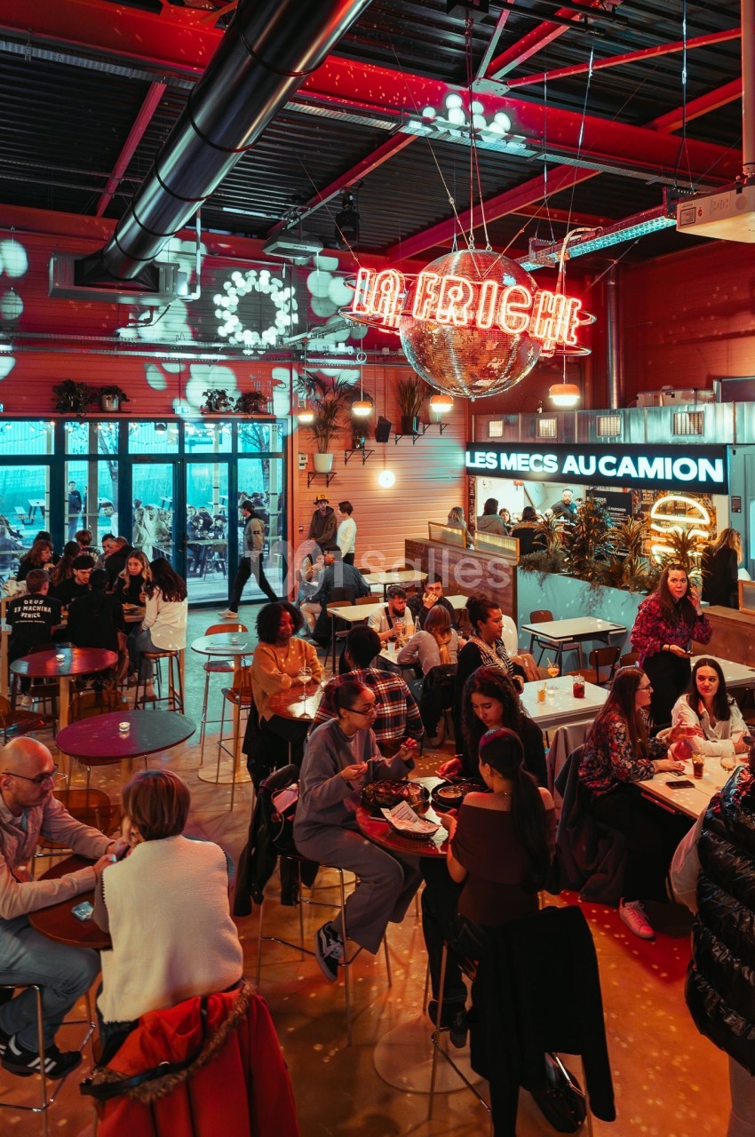 Salle animée avec des groupes de personnes assises à des tables, éclairage coloré et enseignes lumineuses au mur.