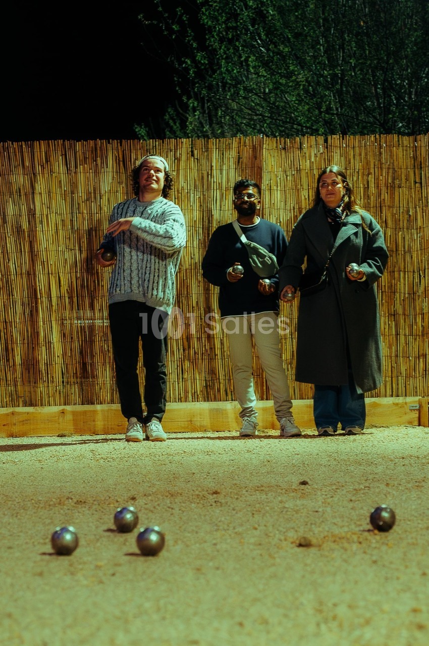 Trois personnes jouent à la pétanque sur un terrain en sable, avec des boules dispersées au sol.