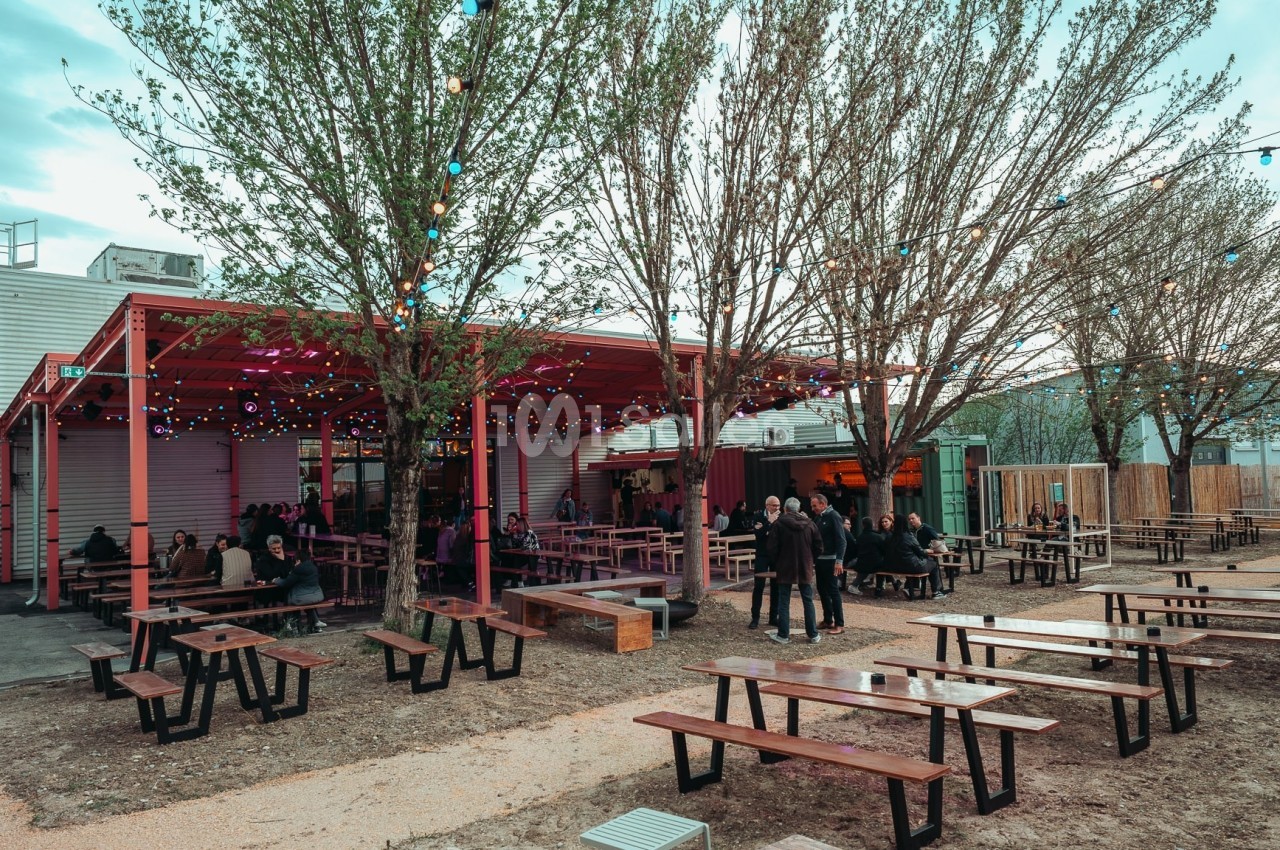 Terrasse extérieure avec tables en bois, éclairée par des guirlandes lumineuses, entourée d'arbres et de bâtiments.