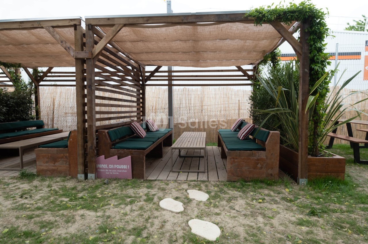 Espace extérieur avec pergolas en bois, banquettes vertes, table basse et végétation environnante.