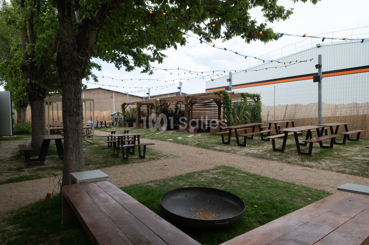 Espace extérieur avec tables de pique-nique, bancs en bois, guirlandes lumineuses et végétation sous une pergola.