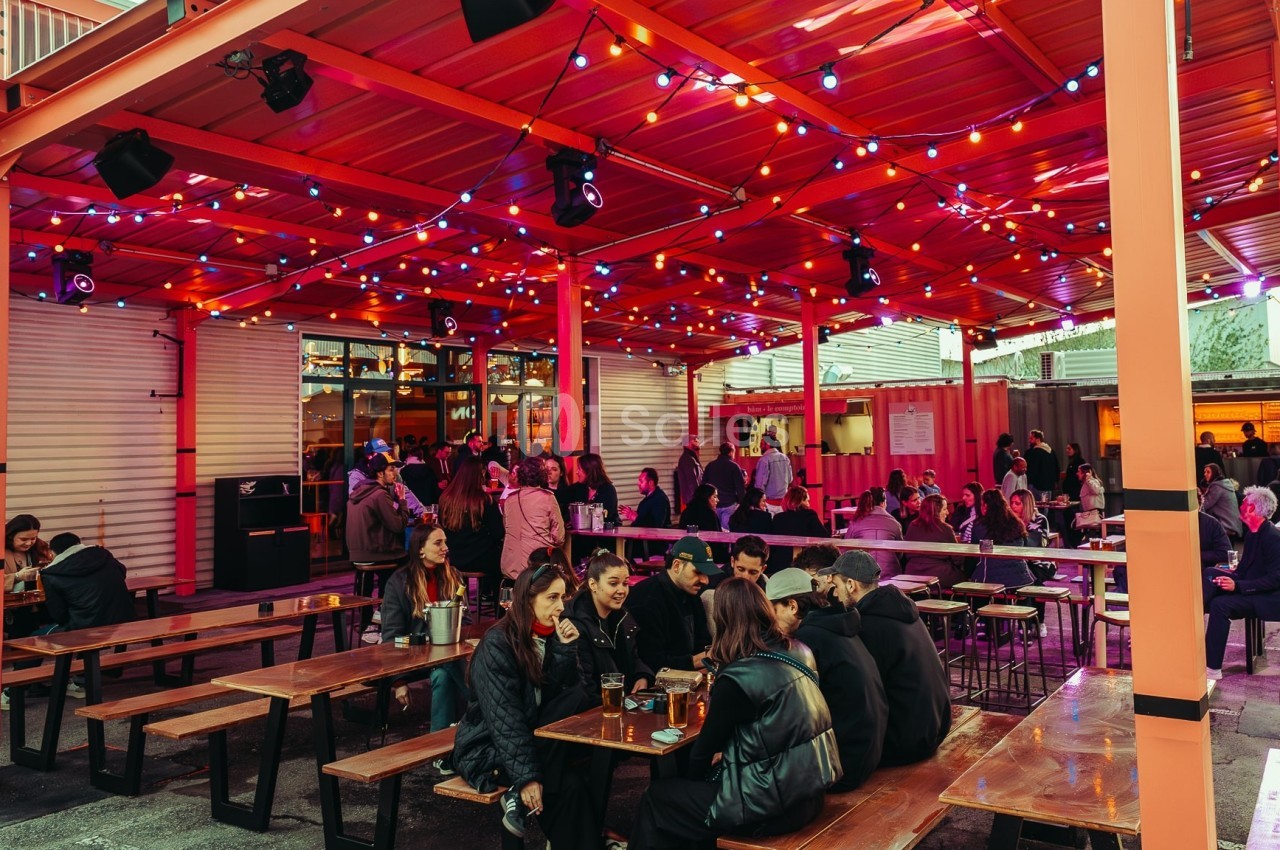 Terrasse extérieure animée avec tables en bois, personnes discutant et éclairage coloré suspendu.