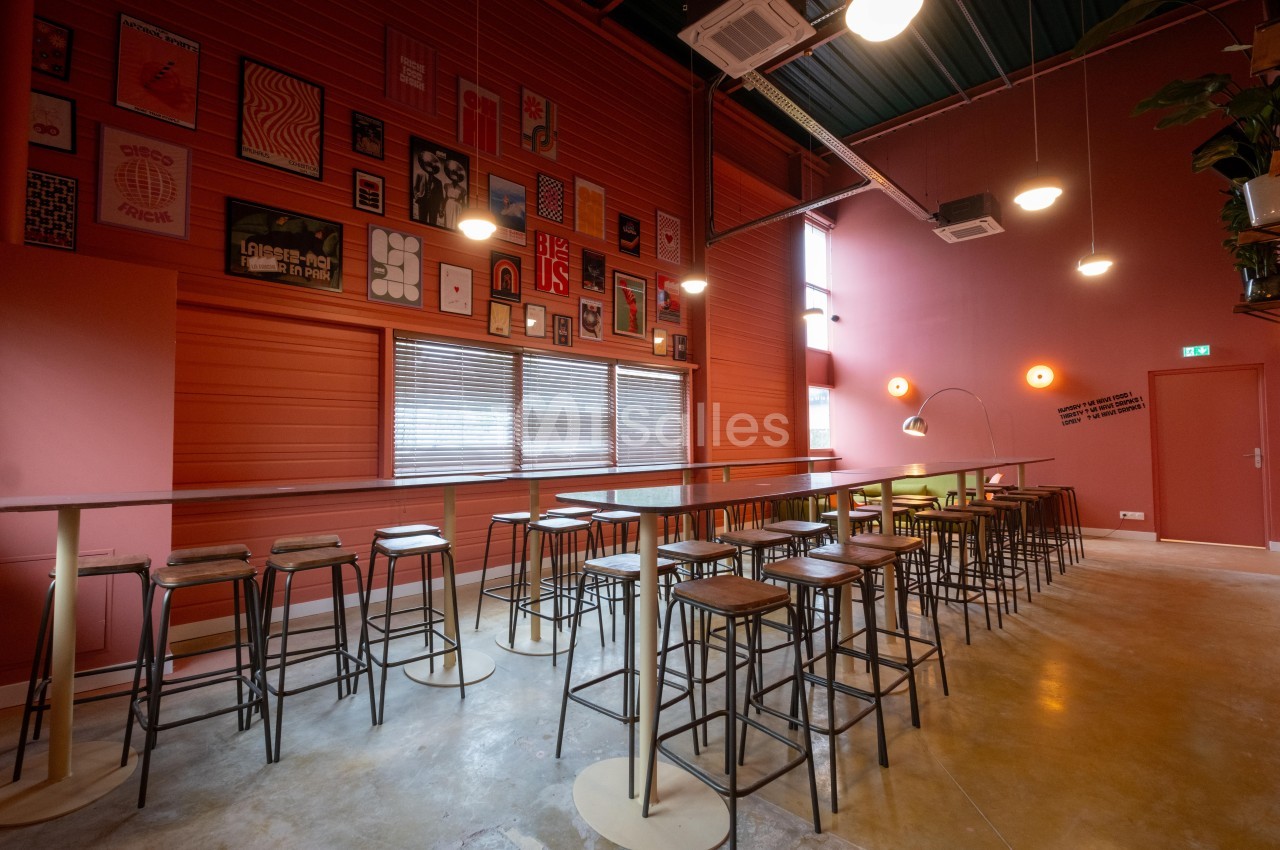 Salle lumineuse avec murs rouges, tables hautes en bois et métal, tabourets, affiches décoratives et luminaires modernes.