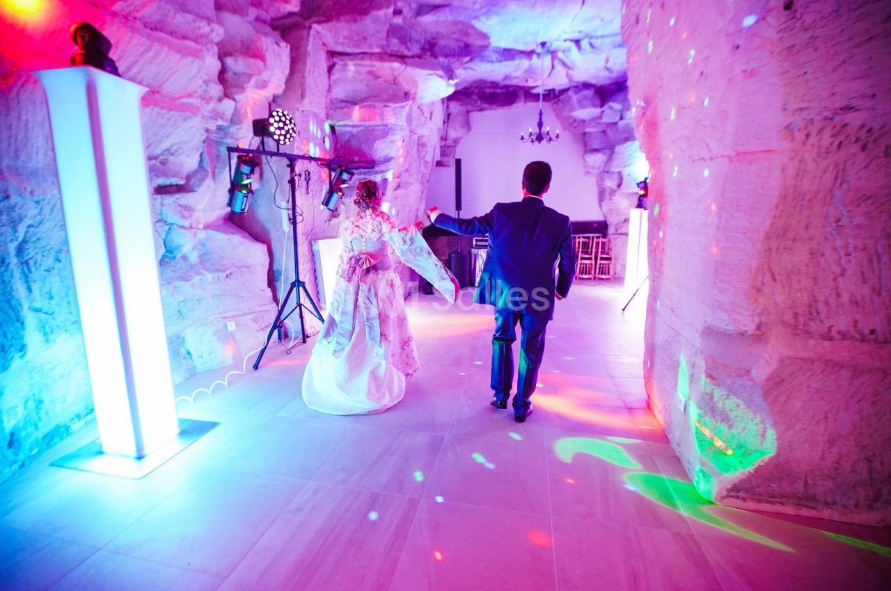 Un couple en tenue de mariage danse dans une salle aux murs de pierre éclairée par des lumières colorées.