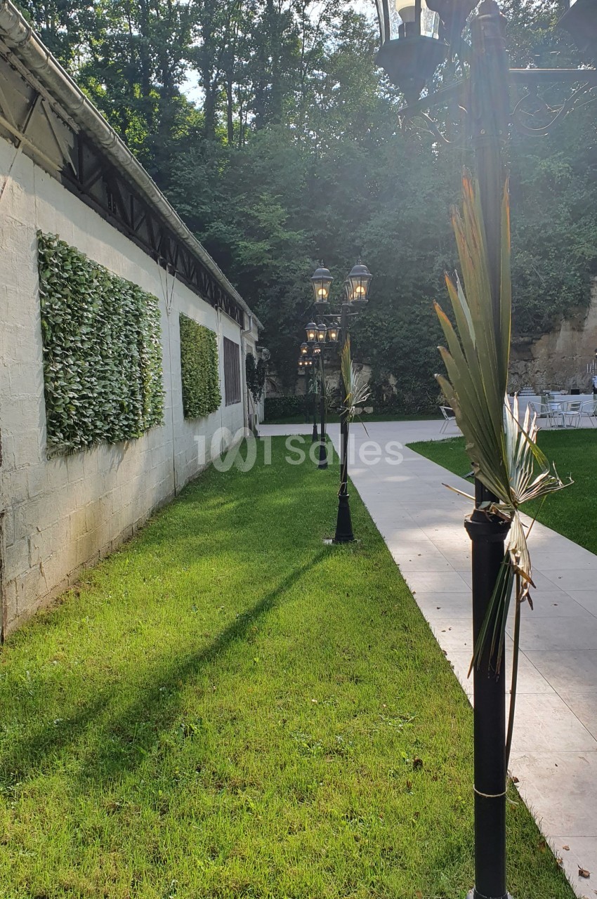 Allée bordée de lampadaires décorés de feuilles, longeant un mur végétalisé et un espace gazonné.