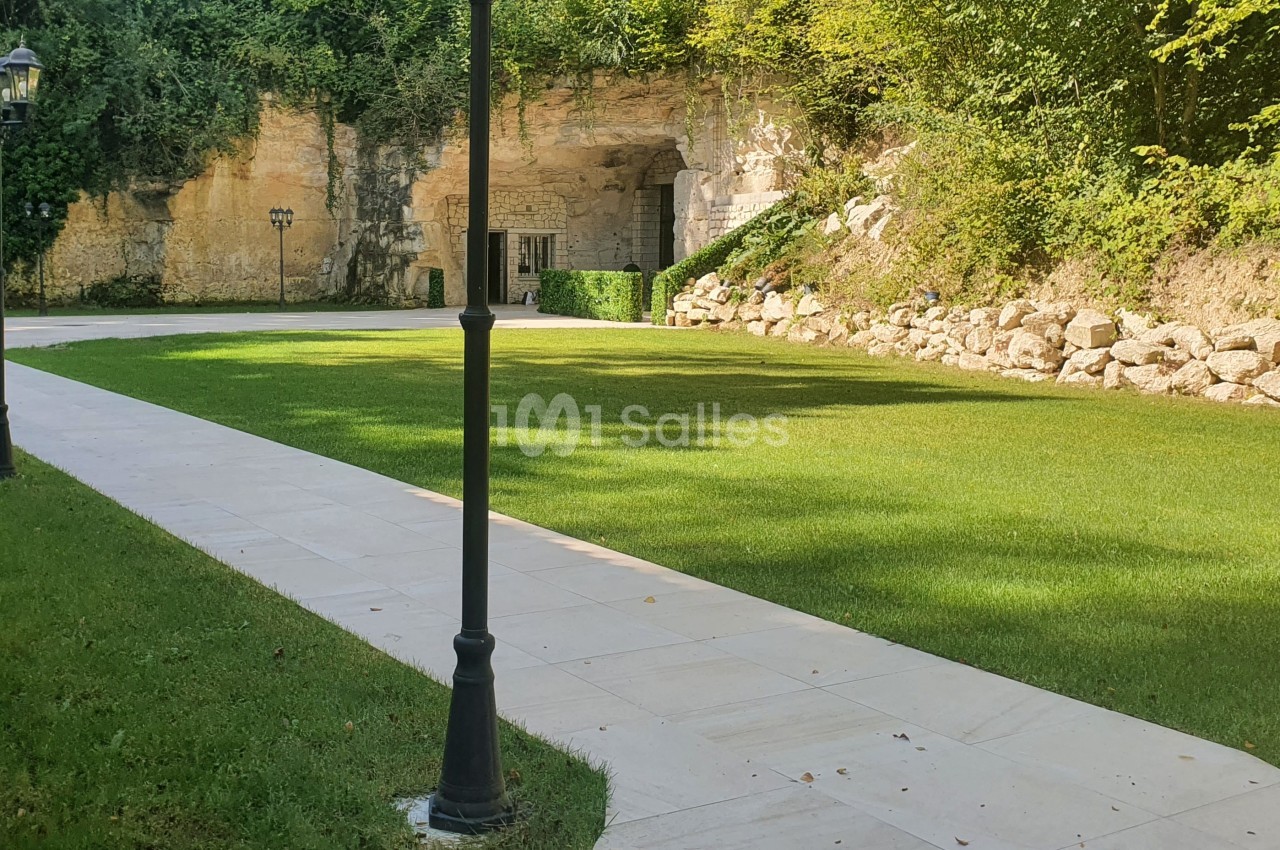 Allée pavée bordée de pelouse menant à une entrée creusée dans une paroi rocheuse, entourée de végétation.