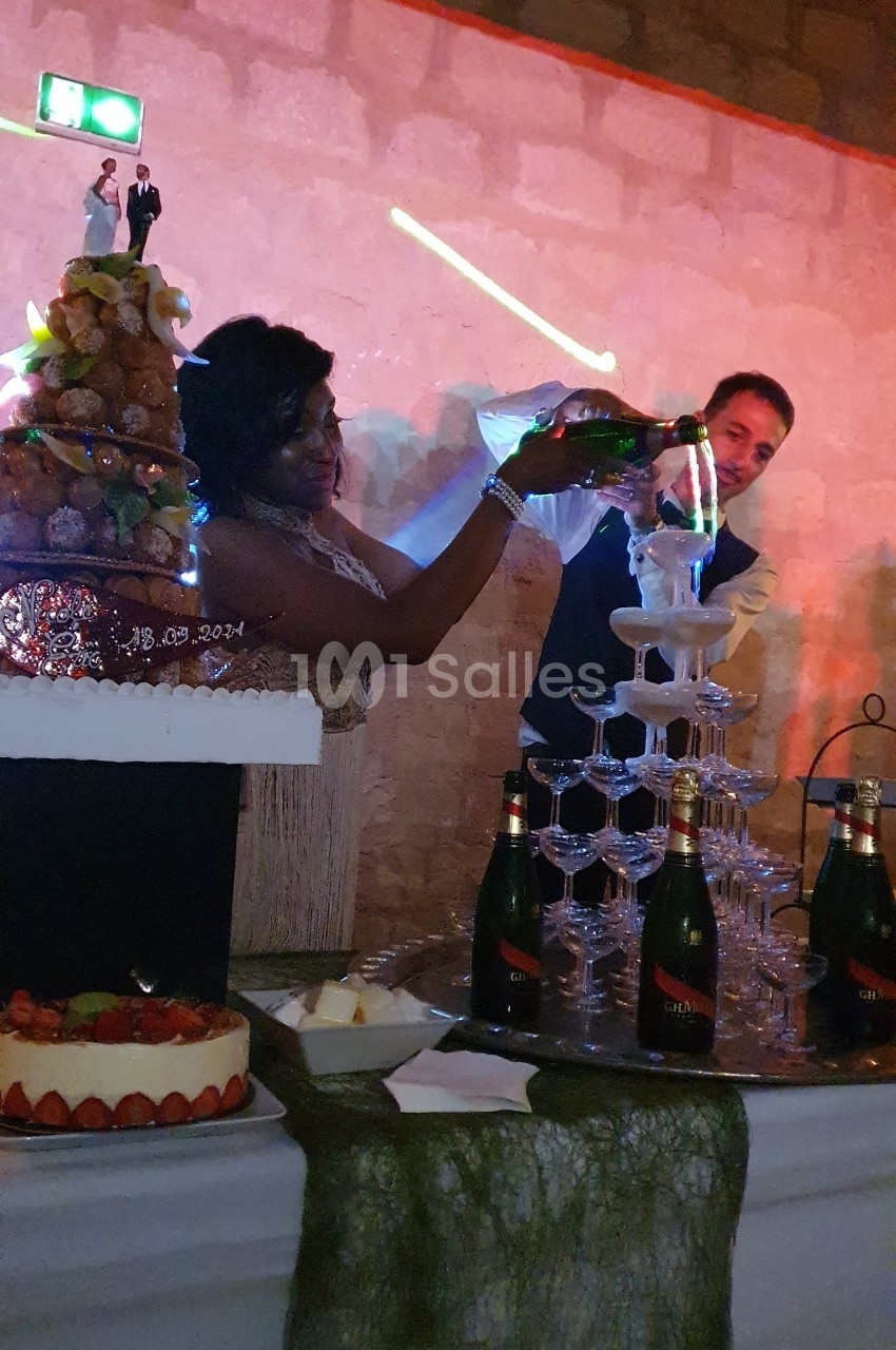 Une femme verse du champagne dans une pyramide de verres lors d'une réception festive, avec un gâteau en arrière-plan.