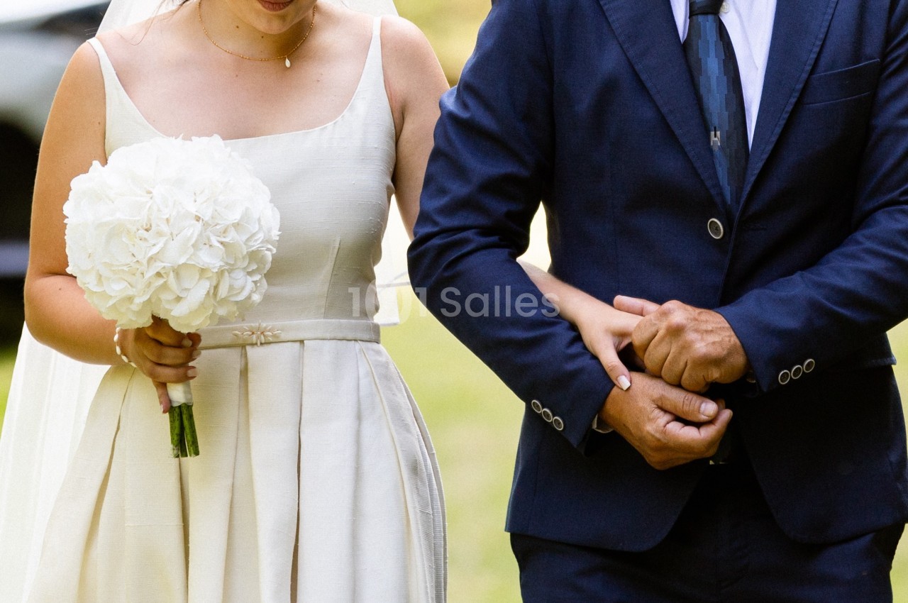 Une mariée en robe blanche tient un bouquet de fleurs blanches, accompagnée d'une personne en costume bleu.