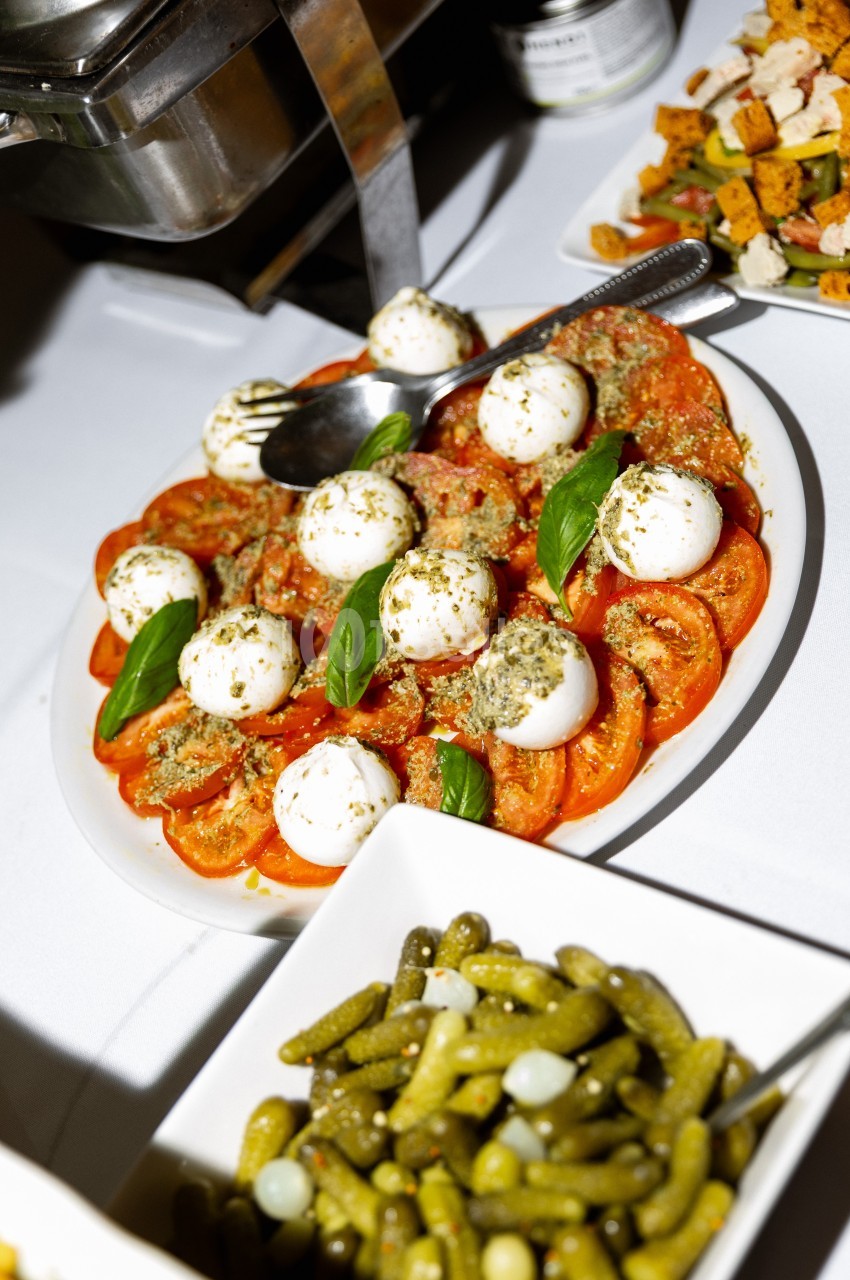 Assiette de tomates, mozzarella, pesto et basilic, accompagnée de cornichons en bocal sur une table de buffet.