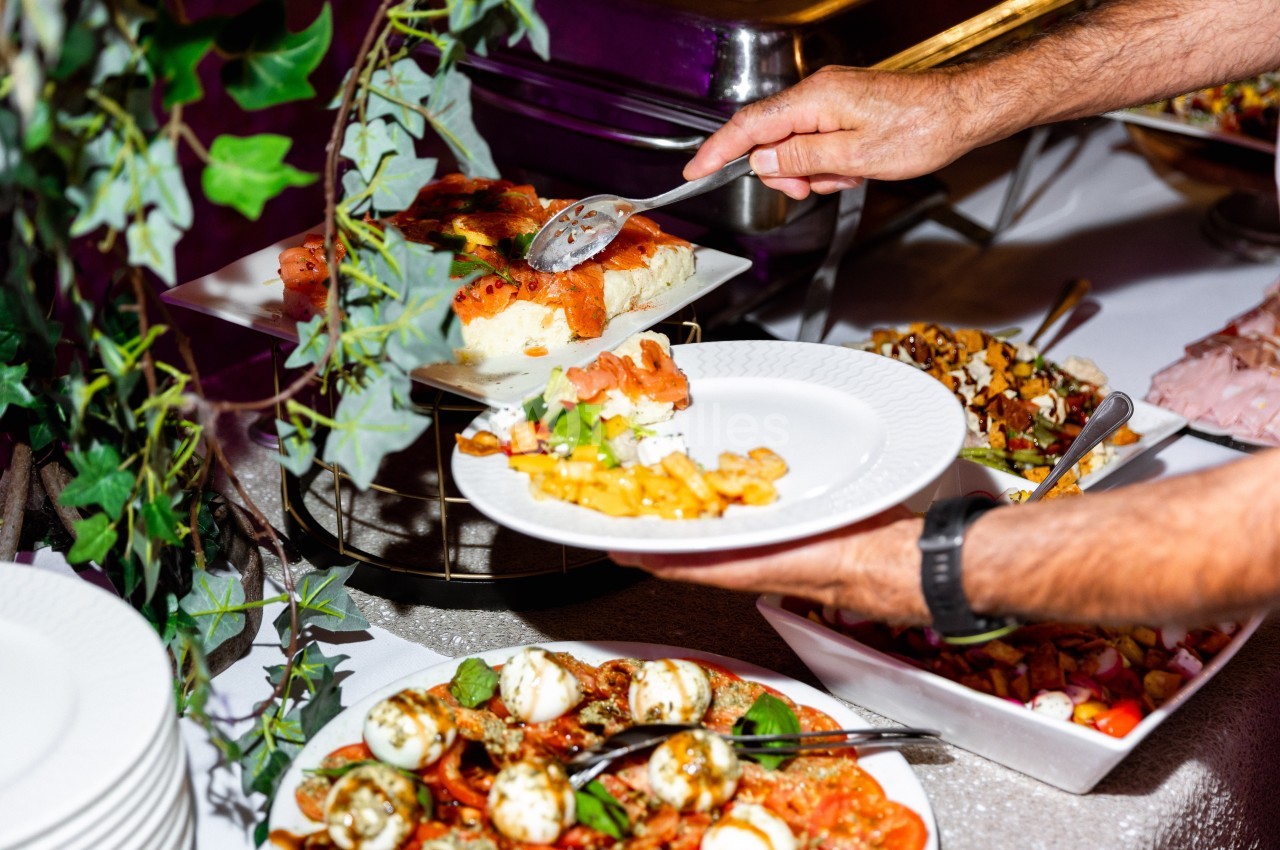 Une personne sert un plat de buffet composé de légumes, salades et autres mets variés sur une assiette blanche.