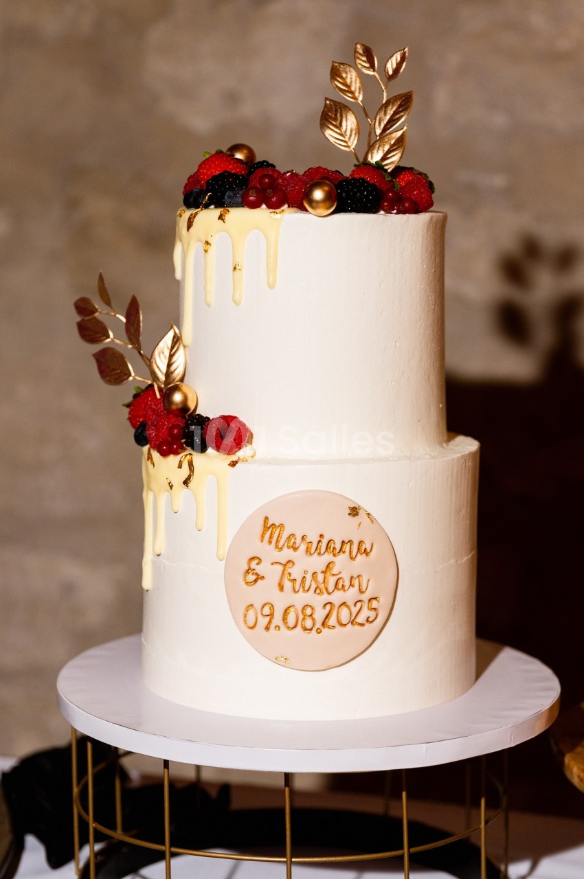 Gâteau de mariage à deux étages décoré de fruits rouges, feuilles dorées et glaçage coulant.