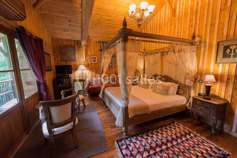 Chambre en bois avec lit à baldaquin, tapis coloré, fauteuils, éclairage chaleureux et vue sur l'extérieur.