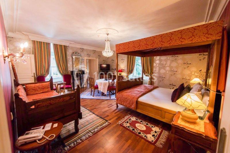 Chambre spacieuse avec lit à baldaquin, coin salon, parquet, tapis et décoration classique.