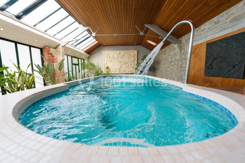 Piscine intérieure avec jets d'eau, entourée de plantes et de murs en pierre, sous un plafond en bois et verrière.