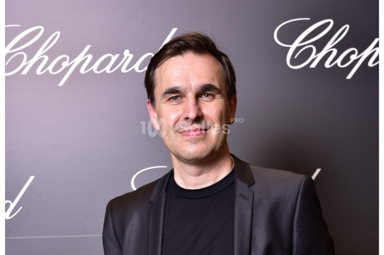 Homme souriant en costume noir devant un fond sombre avec le logo ’Chopard’ répété.