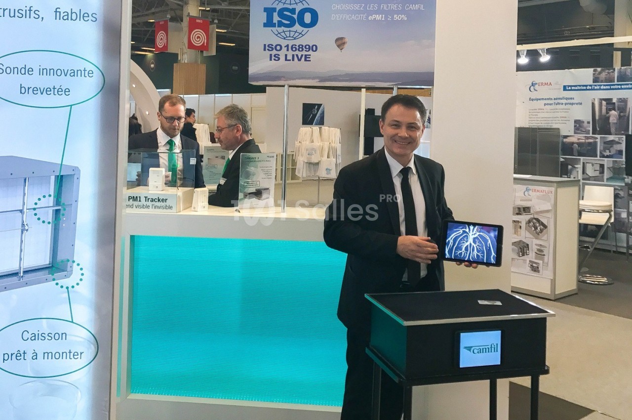 Un homme en costume présente un appareil sur un stand d'exposition, avec des affiches techniques en arrière-plan.