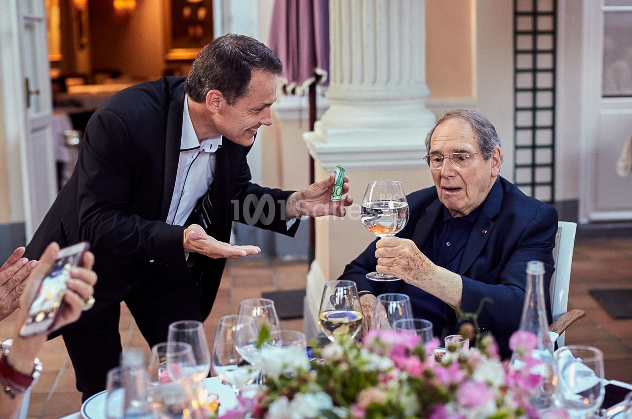Un homme âgé assis lève un verre d'eau, tandis qu'un autre homme debout lui tend un petit objet vert.