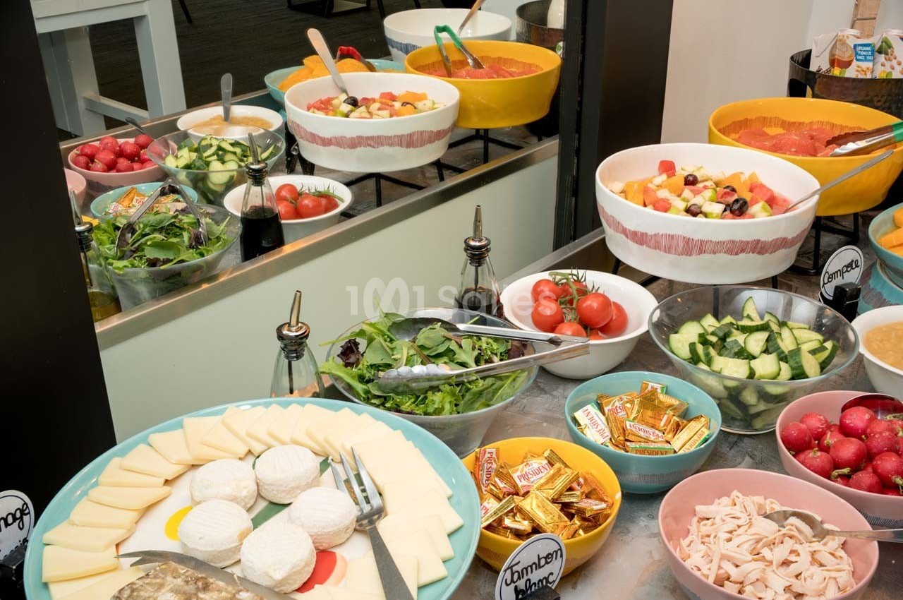 Buffet avec salades, légumes frais, fromages, charcuterie et condiments disposés dans des bols colorés.
