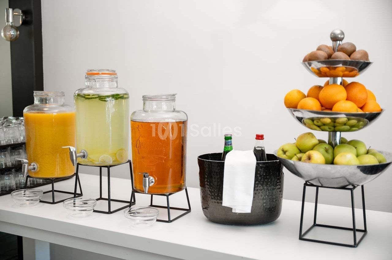 Distributeurs de jus et d'eau citronnée, fruits frais sur un présentoir et boissons dans un seau sur une table blanche.
