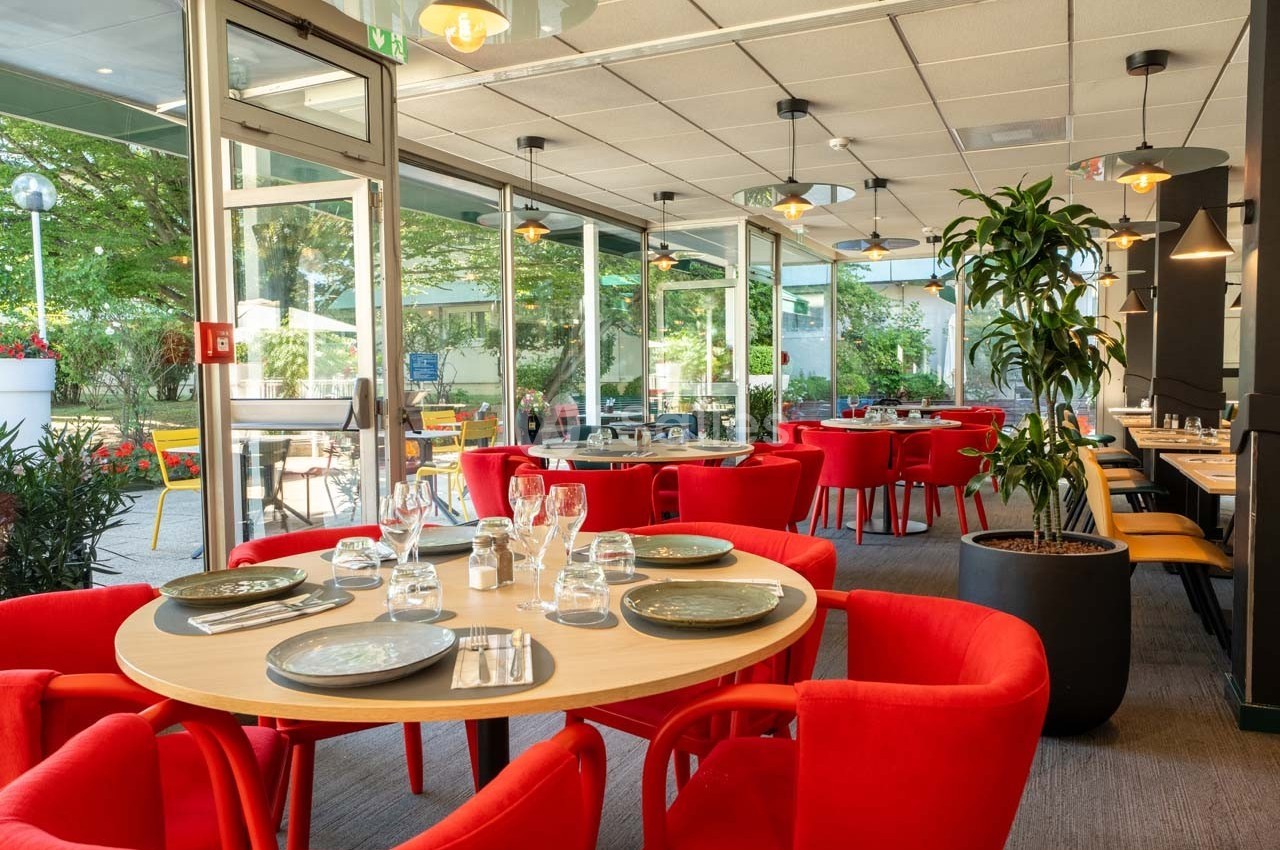 Salle de restaurant lumineuse avec des tables dressées, chaises rouges et vue sur une terrasse arborée.