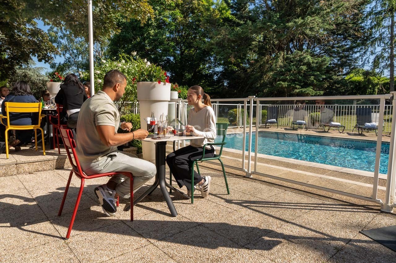 Deux personnes déjeunent en terrasse près d'une piscine, entourées de chaises colorées et d'arbres.