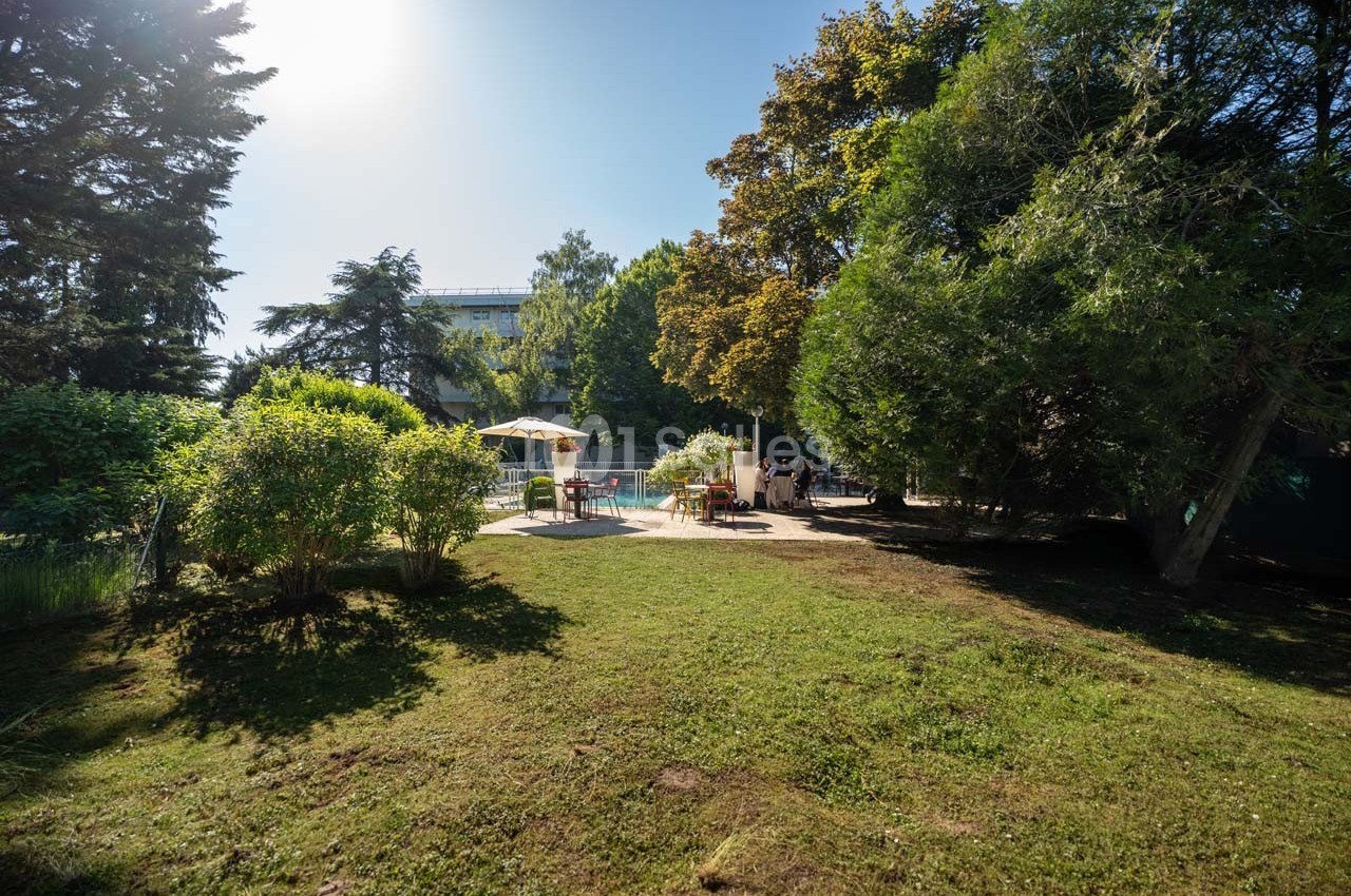 Espace extérieur verdoyant avec pelouse, arbres et une piscine entourée de chaises et parasols sous un ciel ensoleillé.