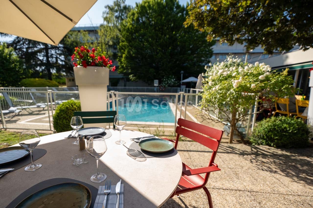 Table dressée en extérieur près d'une piscine, entourée de verdure et de fleurs sous un parasol.