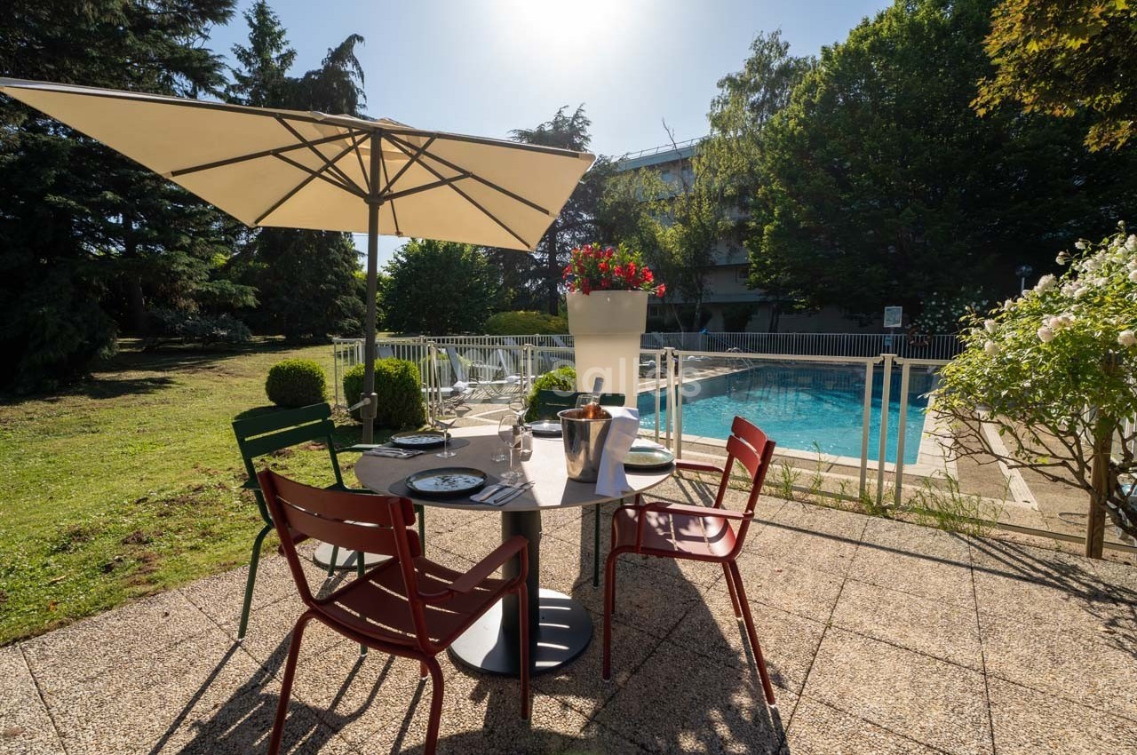 Table dressée en extérieur avec parasol, vue sur une piscine entourée de verdure et baignée de lumière.