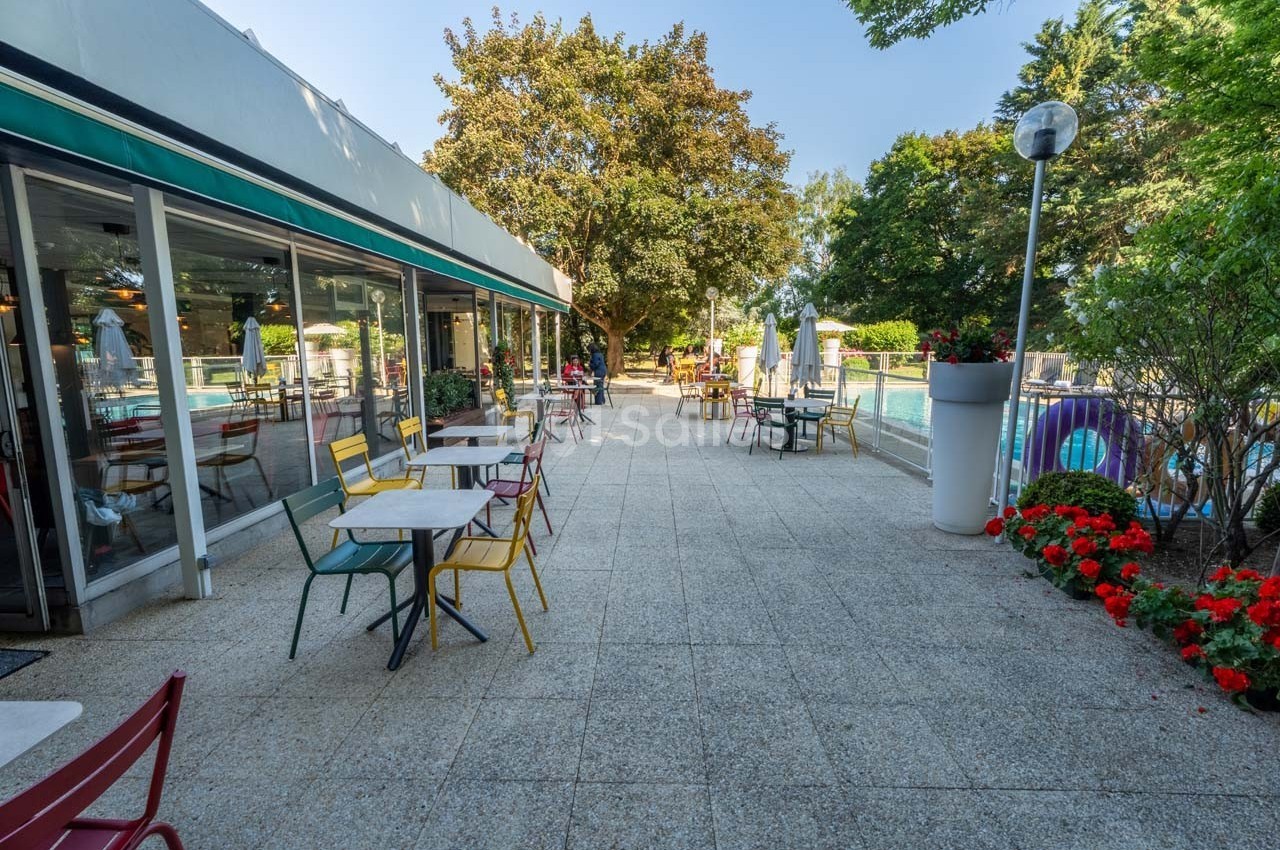 Terrasse extérieure avec tables et chaises colorées, bordée de fleurs et donnant sur une piscine entourée d'arbres.