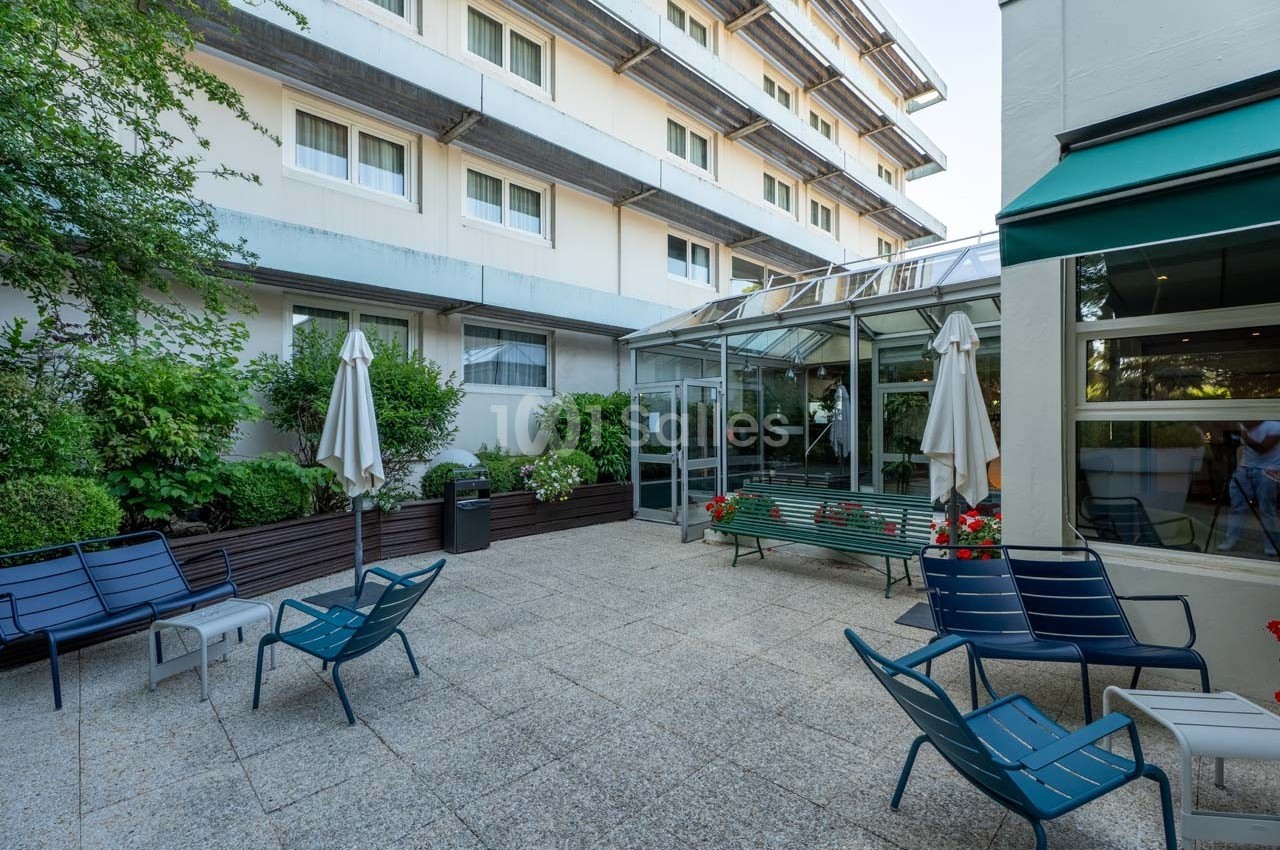 Cour extérieure avec mobilier de jardin, parasols fermés, plantes et accès vitré à un bâtiment moderne.