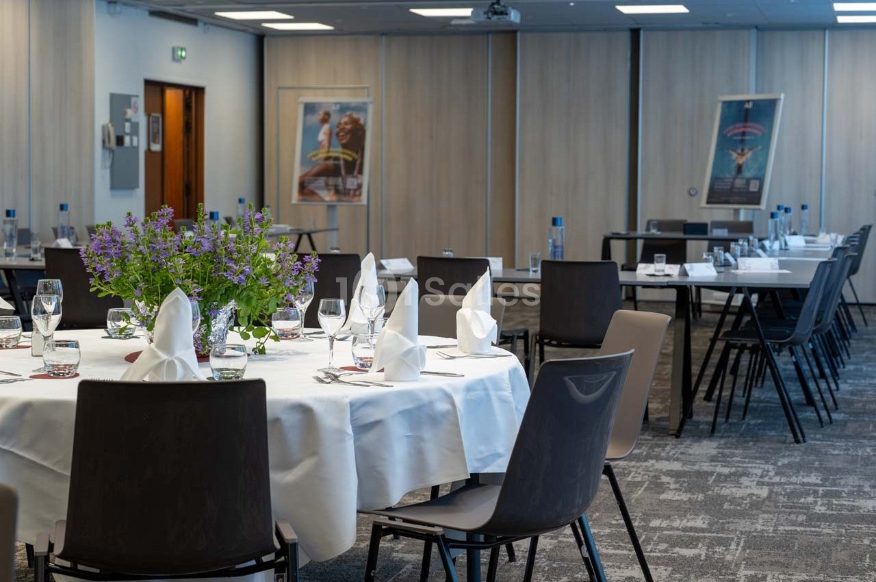 Salle de réunion aménagée avec des tables rondes dressées, décorées de fleurs et entourées de chaises.