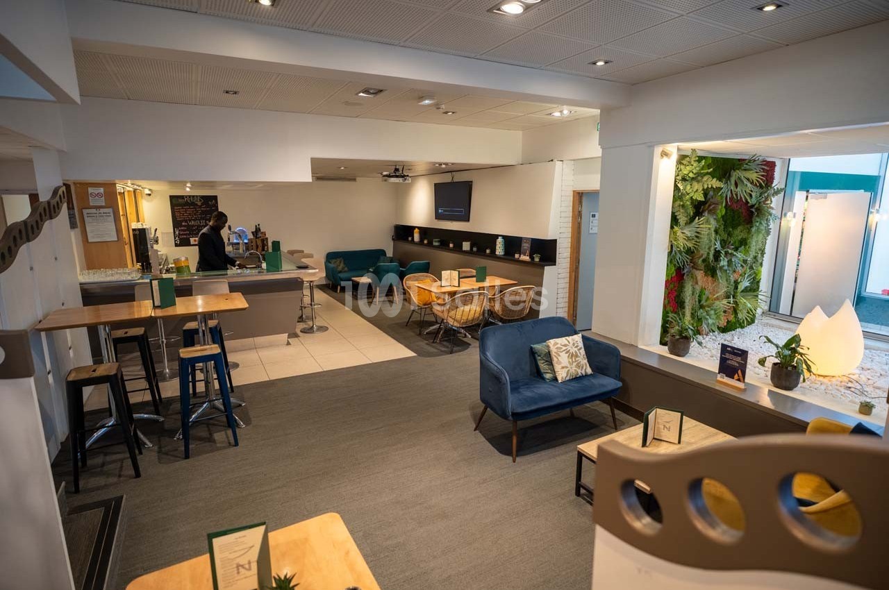 Espace lounge lumineux avec tables, chaises, fauteuils et mur végétal, aménagé pour détente ou travail.