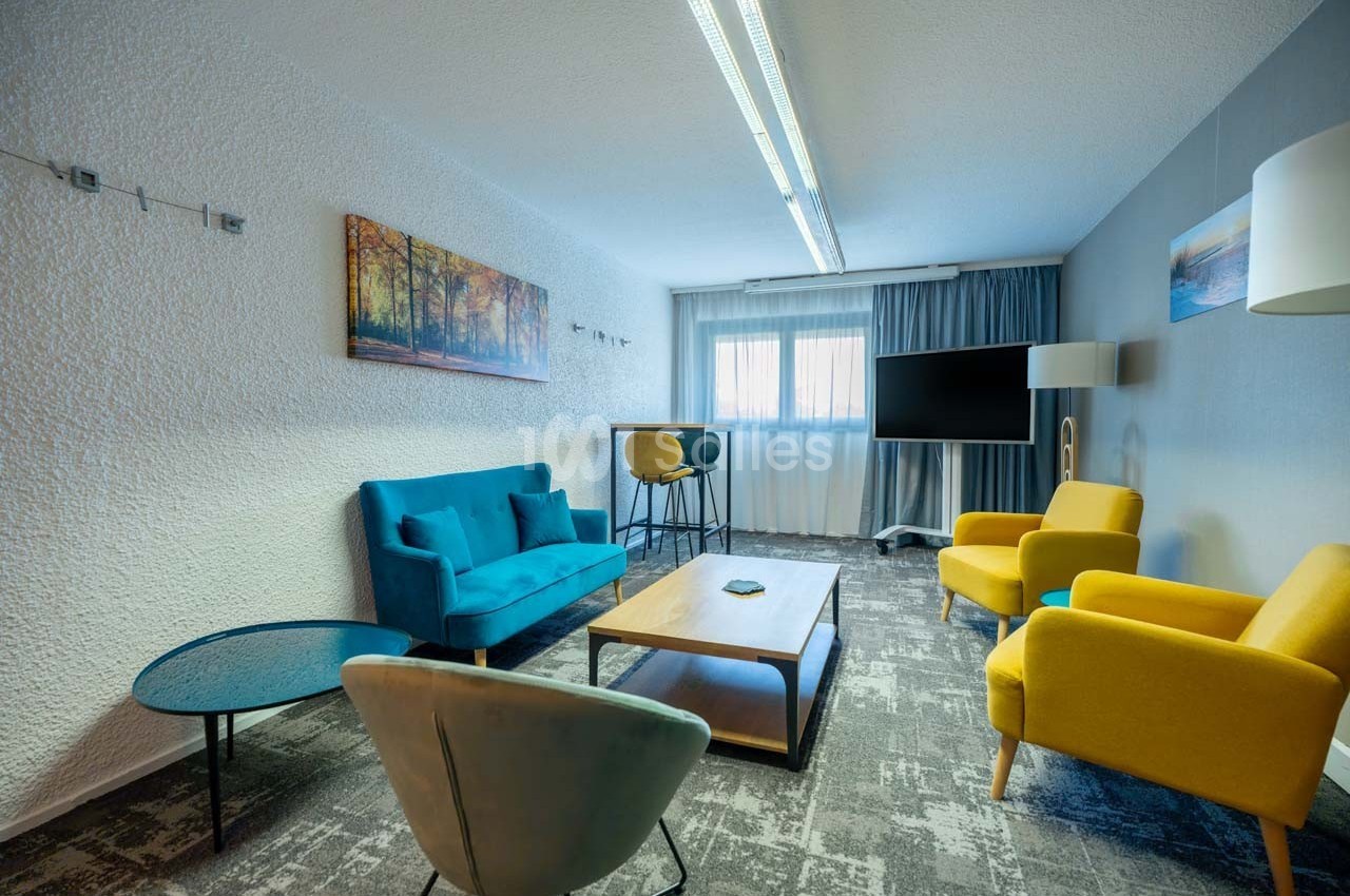 Salle de réunion lumineuse avec canapé bleu, fauteuils jaunes, table basse en bois et écran de télévision.