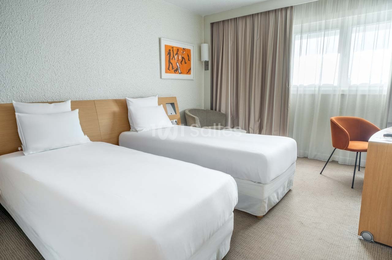 Chambre d'hôtel avec deux lits simples, un bureau, une chaise orange et une grande fenêtre avec rideaux beige.