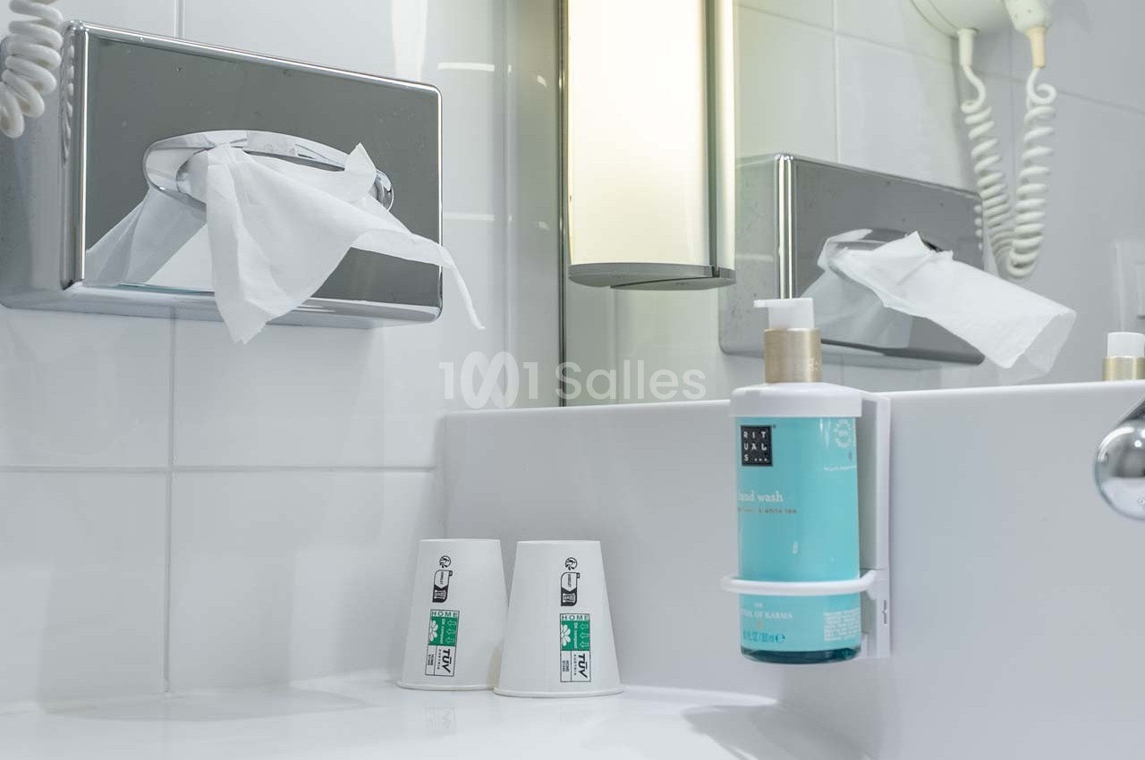 Distributeur de mouchoirs, savon liquide et gobelets en plastique sur un lavabo dans une salle de bain lumineuse.