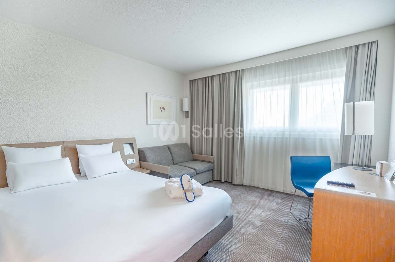 Chambre d'hôtel lumineuse avec lit double, canapé, bureau, chaise bleue et grande fenêtre avec rideaux.