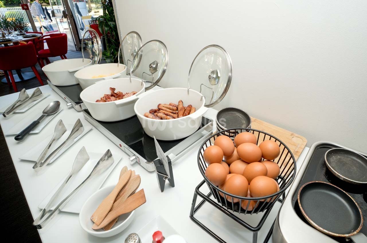 Buffet de petit-déjeuner avec œufs, saucisses, bacon et ustensiles disposés sur un comptoir blanc.