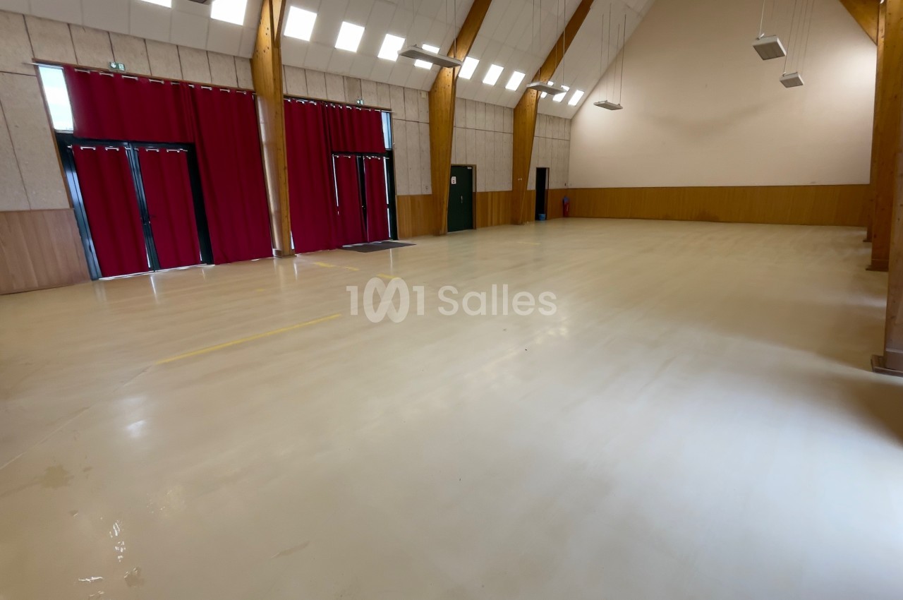 Salle vide avec un sol clair, des murs en bois, des poutres apparentes et des rideaux rouges couvrant des ouvertures.