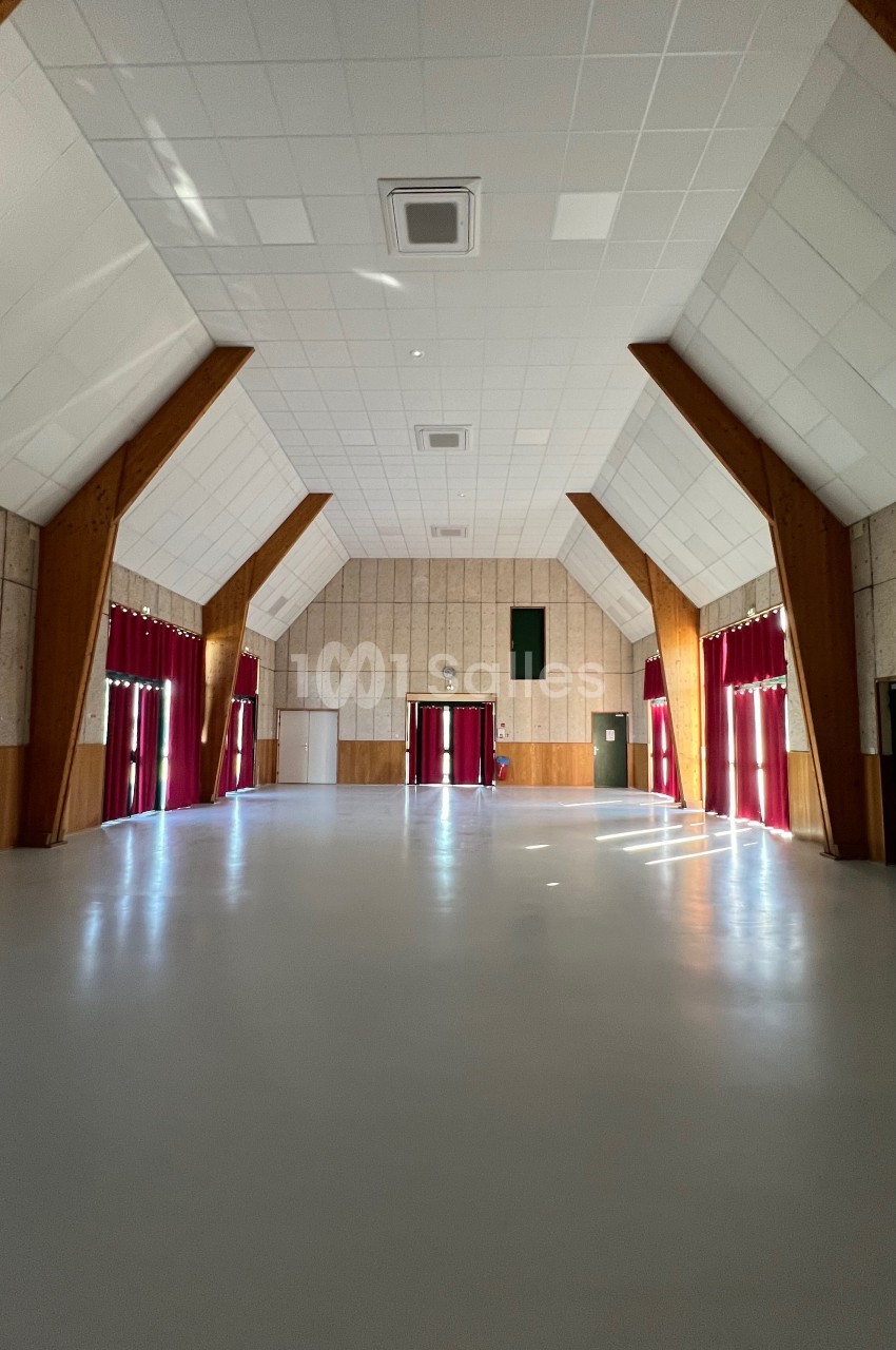 Salle vide avec un sol clair, des murs en bois et des rideaux rouges encadrant de grandes fenêtres.