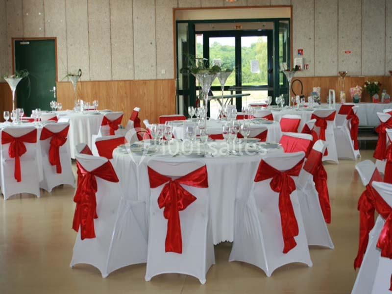 Salle décorée pour un événement avec des tables rondes, nappes blanches et chaises ornées de nœuds rouges.