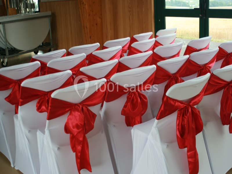 Chaises blanches alignées, décorées de nœuds rouges en tissu dans une salle avec des murs en bois et une baie vitrée.