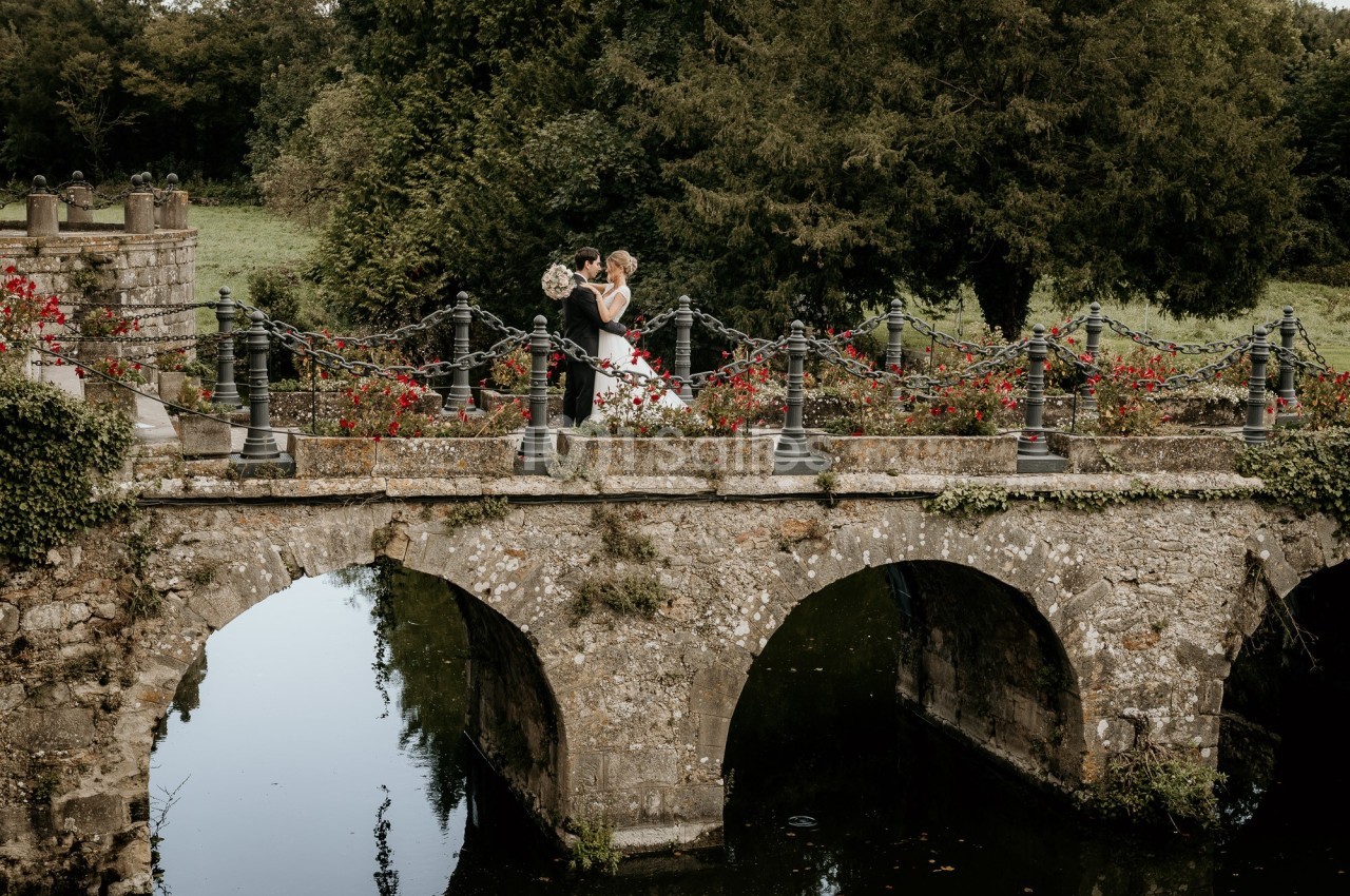 Un couple en tenue de mariage se tient sur un pont en pierre entouré de fleurs et de verdure.